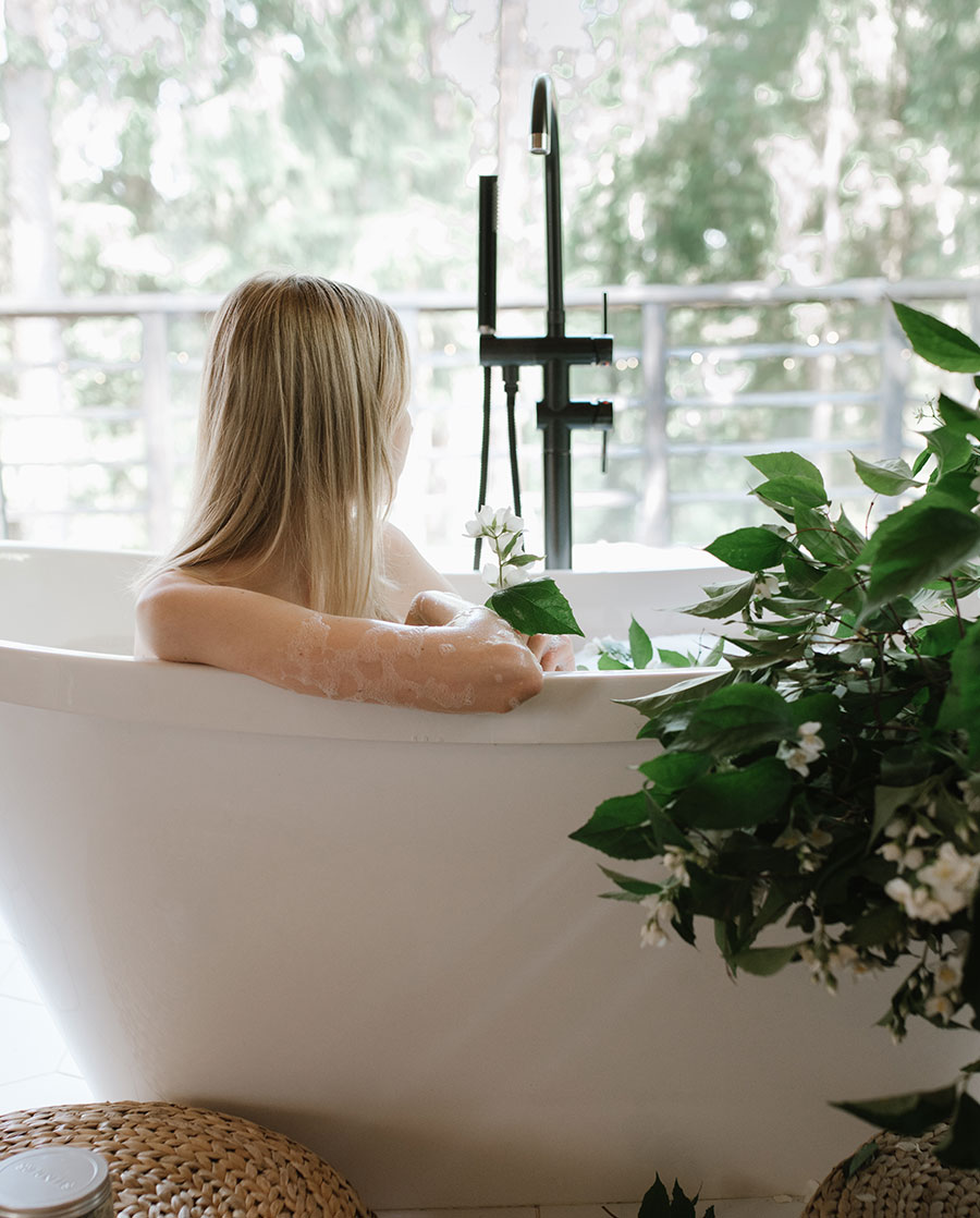 A woman resting in a freestanding bath beside blossoming jasmine and a forest window